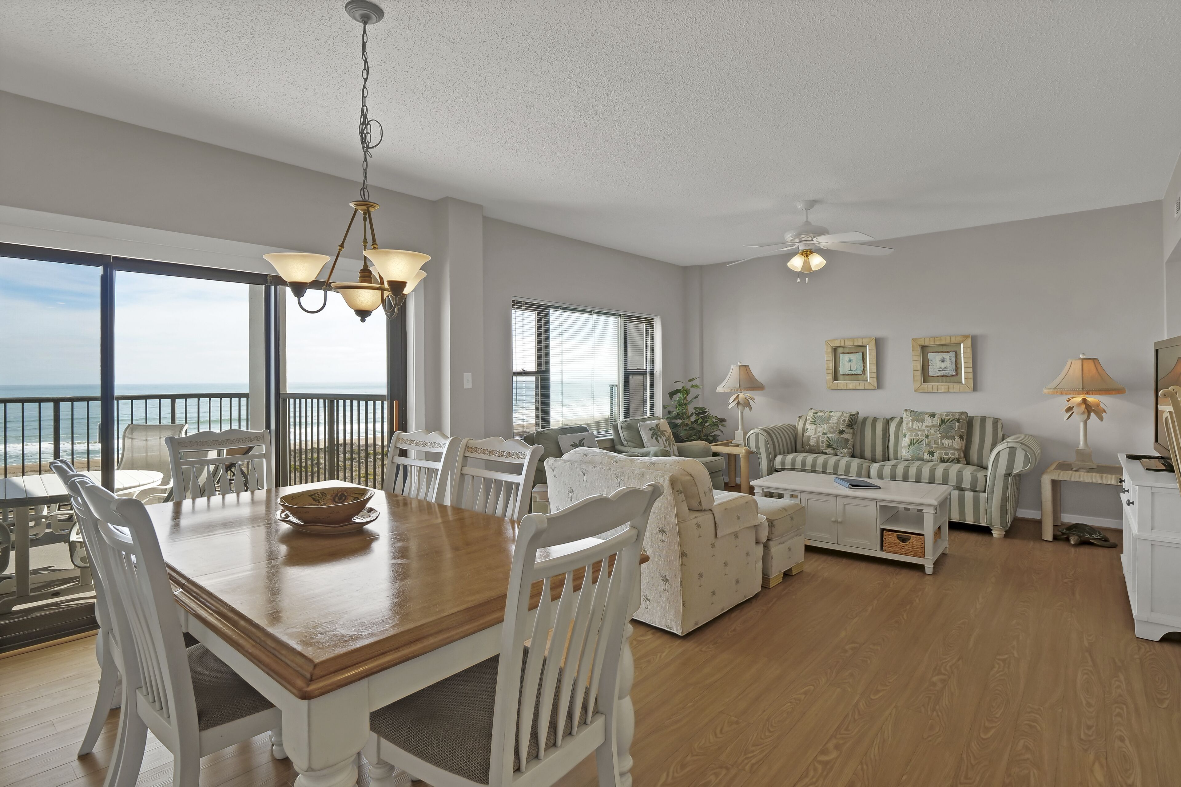 A spacious living room with a wooden dining table, white chairs, a sofa, and a ceiling fan.