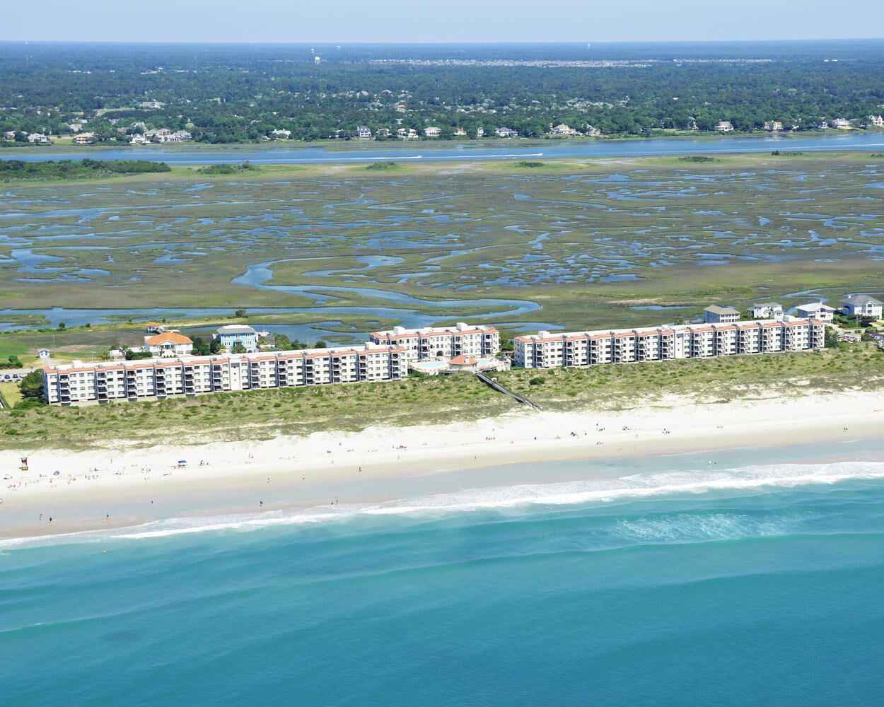 Un hôtel en bord de mer, composé de plusieurs bâtiments, avec une plage de sable et une zone marécageuse en arrière-plan.