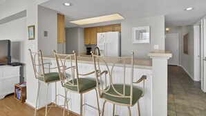 A kitchen with wooden cabinets, a white refrigerator, and a bar area with two chairs. - DR 1405 - Beautiful oceanfront condo with tennis, pool and easy beach access (Wrightsville Beach)