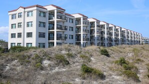 Un immeuble résidentiel à plusieurs étages, doté de balcons, situé sur un terrain sablonneux où la végétation est clairsemée.