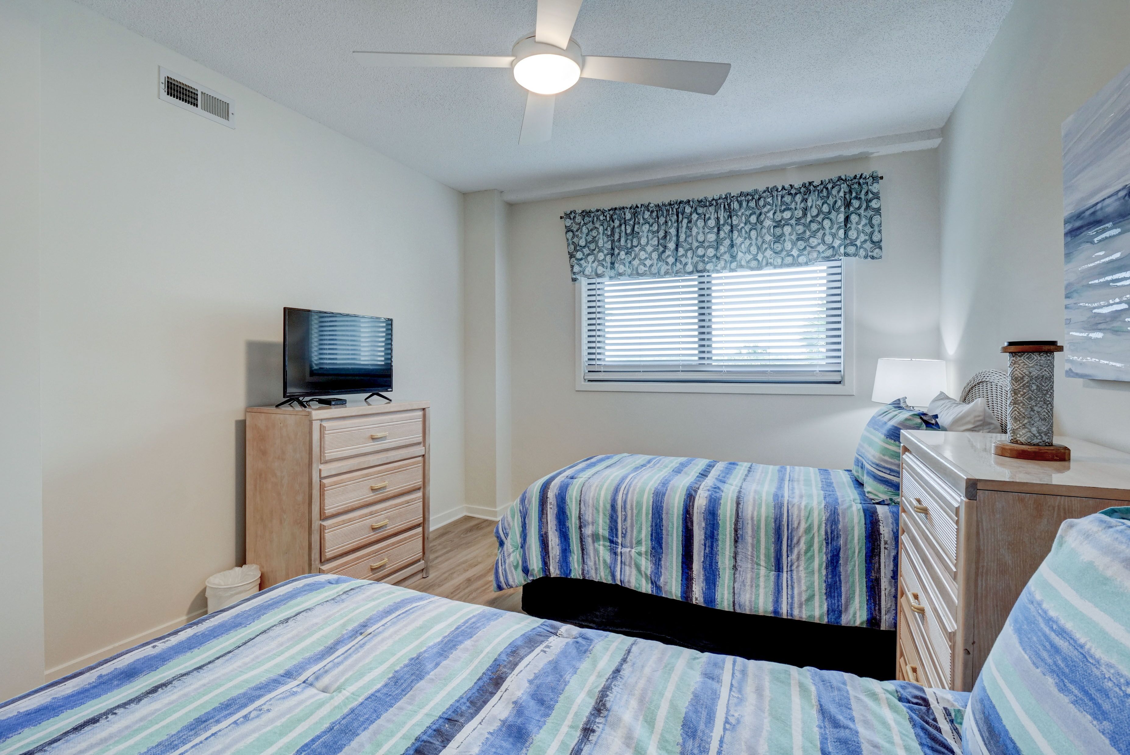 A bedroom with two beds, a ceiling fan, a television on a dresser, and a window with blinds.