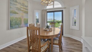 Un comedor con una mesa redonda de madera, seis sillas y una araña. Hay un cuadro grande en la pared y vistas a través del ventanal.