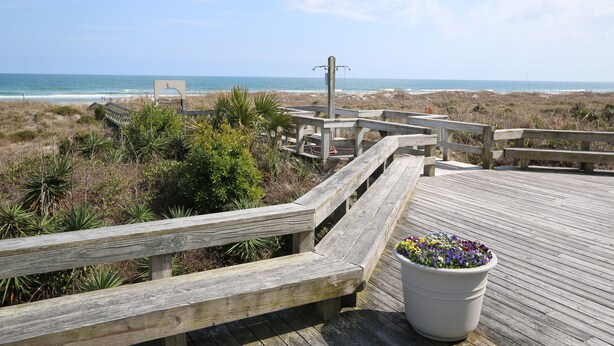 Un paseo marítimo de madera que lleva a la playa, con una banca y una jardinera con flores.