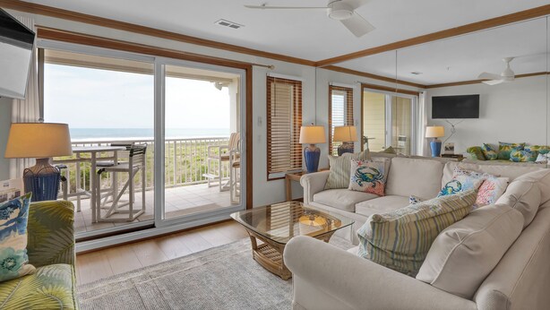 A spacious living room with a glass sliding door leading to a balcony, a ceiling fan, and a flat-screen TV mounted on the wall. - Wrightsville Dunes F-2B - Oceanfront condo with community pool, tennis, beach (Wrightsville Beach)