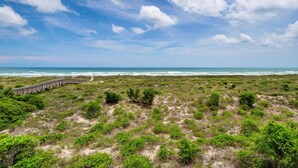 A coastal landscape with a wooden boardwalk leading to the beach, surrounded by green vegetation. - Wrightsville Dunes F-2B - Oceanfront condo with community pool, tennis, beach (Wrightsville Beach)
