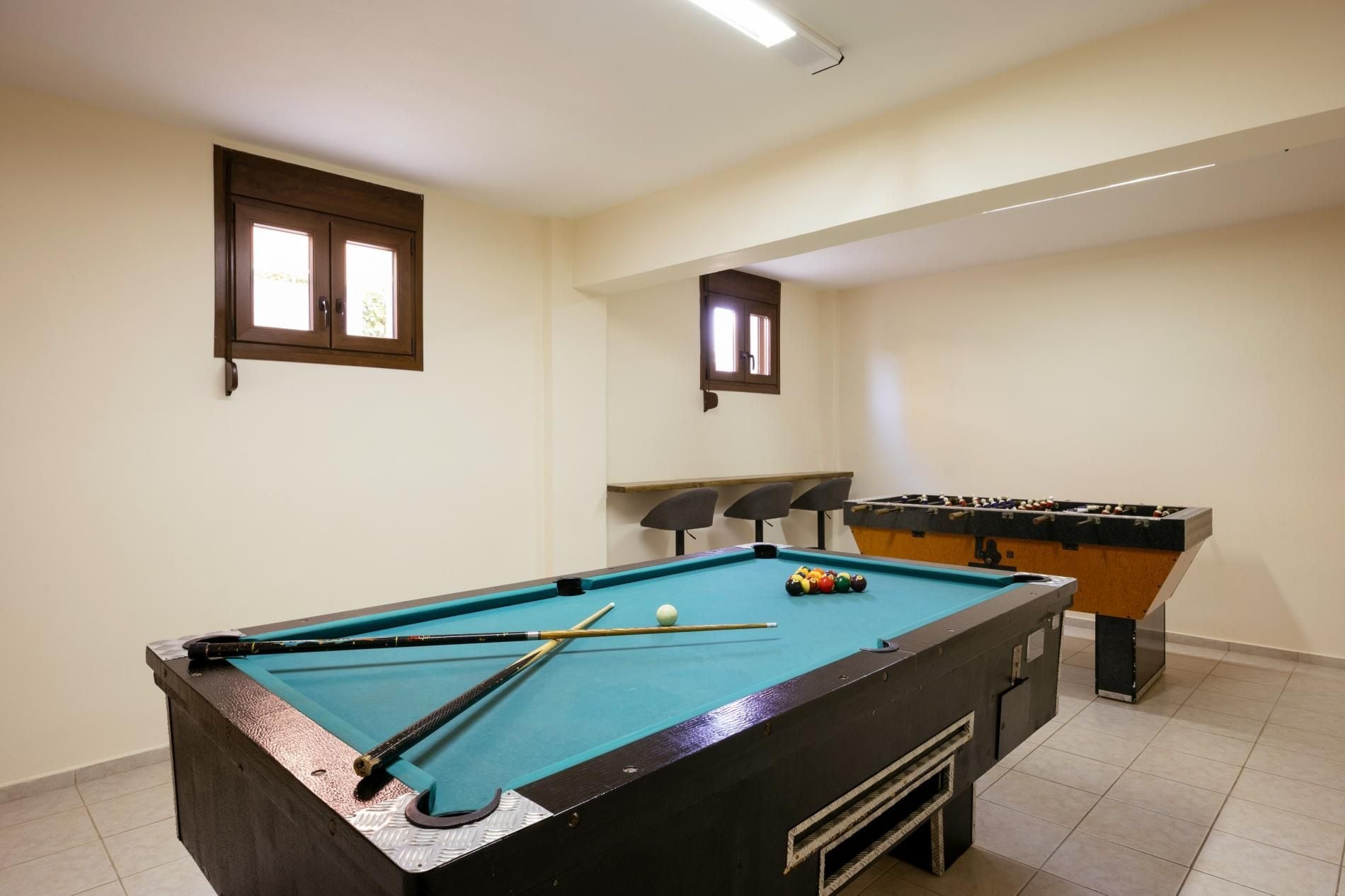 Una mesa de billar con tacos y bolas, una mesa de fútbol de mesa y dos ventanas con persianas.