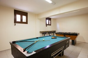 A pool table with cues and balls, a foosball table, and two windows with blinds.