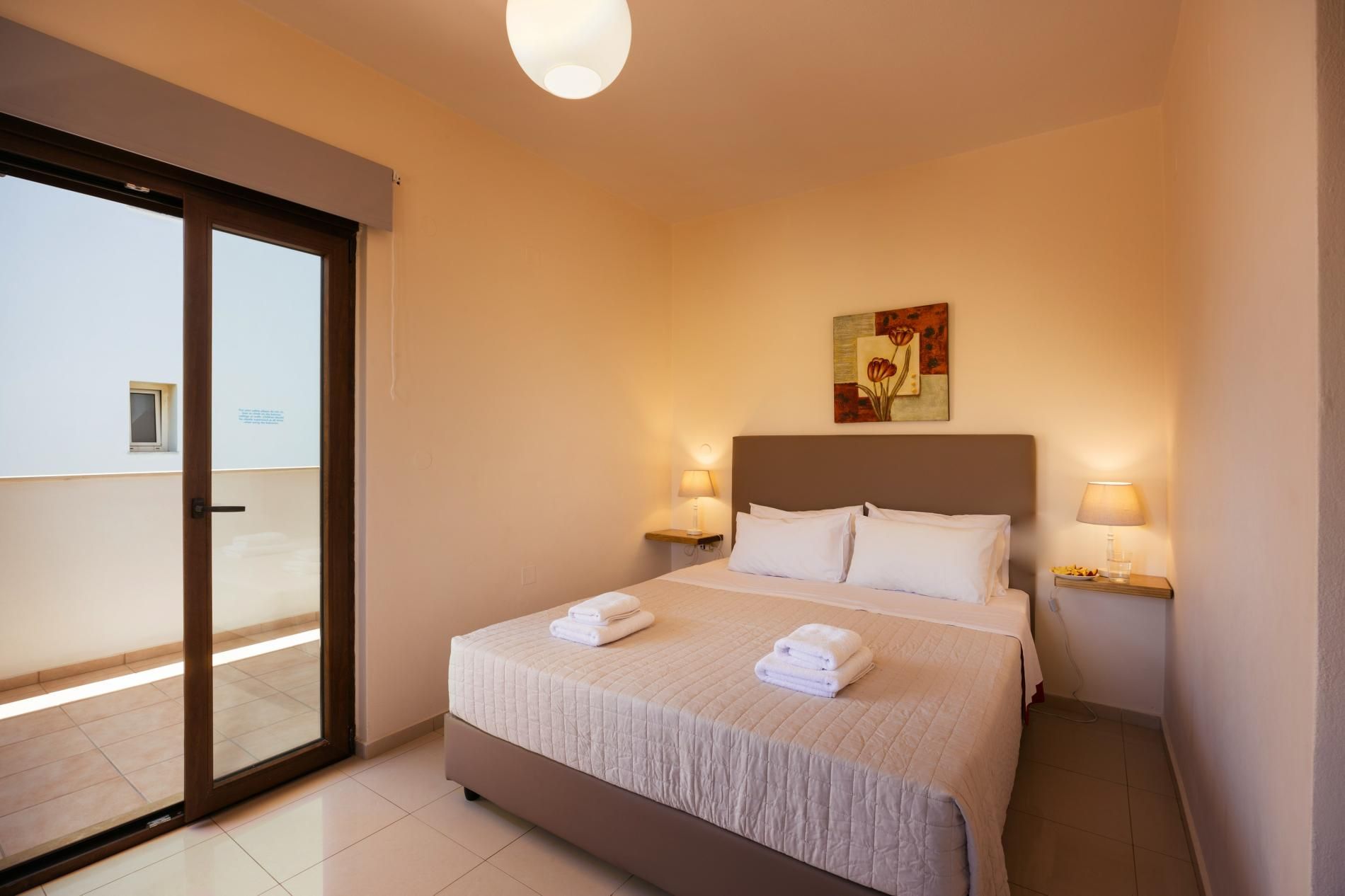 Quarto de hotel com cama, duas luminárias de cabeceira, um quadro na parede e uma porta de vidro deslizante para área externa.