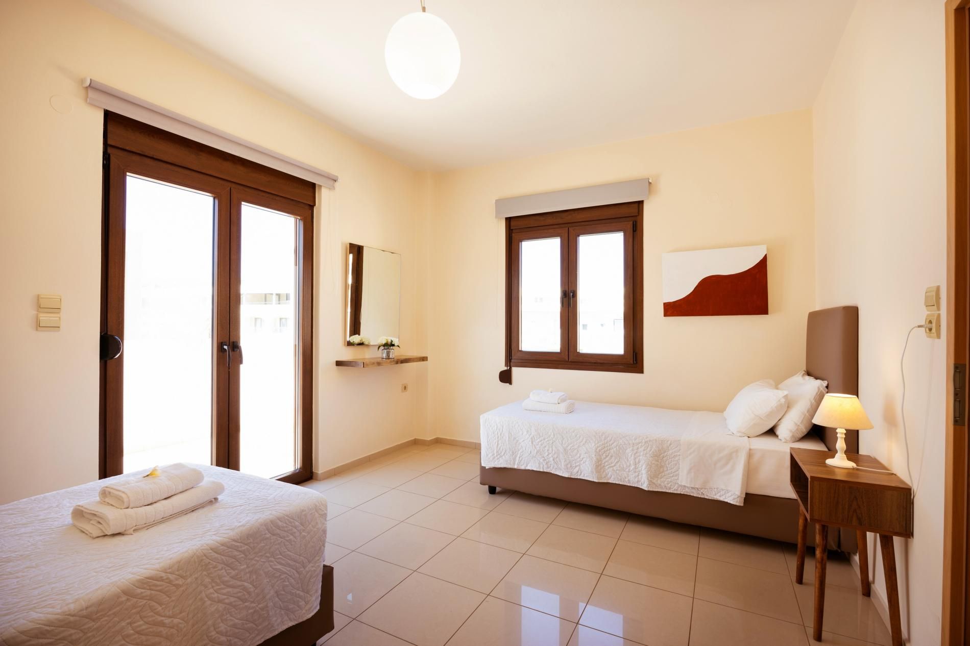 A hotel room with two beds, a bedside table with a lamp, a mirror, and a window with blinds.
