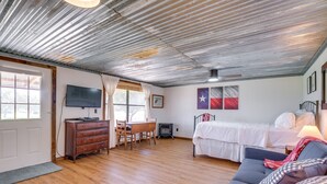 Une pièce avec un plancher en bois, un lit avec une literie blanche, un canapé gris, une commode en bois, un téléviseur et un drapeau accroché au mur.