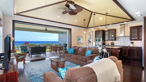 A spacious living area with a large window overlooking the ocean, a ceiling fan, and a kitchen with dark wooden cabinets.