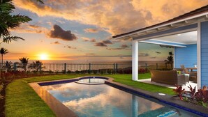 A swimming pool with a view of the sunset over the ocean, a patio area with outdoor furniture, and a blue house with a white exterior.