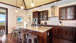 A modern kitchen with dark wood cabinets, a large island with bar stools, and a view of the ocean through the sliding glass door.