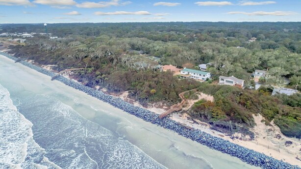 A coastal area with a beach, rocky shoreline, and residential buildings surrounded by dense vegetation. - Beachfront Bliss 4 bedrooms, 3 bathrooms (Jekyll Island)