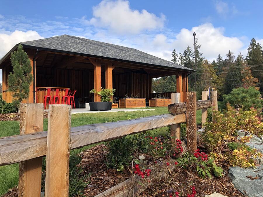 Un bâtiment rustique en bois doté d’un patio couvert, d’un espace extérieur aménagé avec des sièges et d’un jardin où poussent diverses plantes et fleurs.