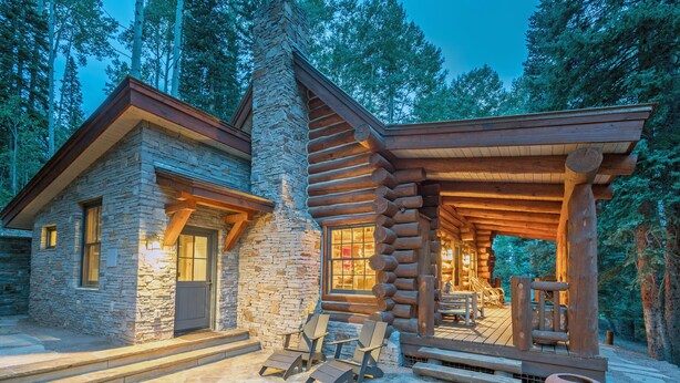 Une cabane en rondins avec des murs en pierre, une véranda avec des chaises en bois et une cheminée en pierre.