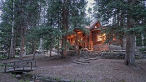 Une cabane en rondins avec un escalier en pierre menant à une terrasse en bois, entourée de grands arbres et d’un petit ruisseau.