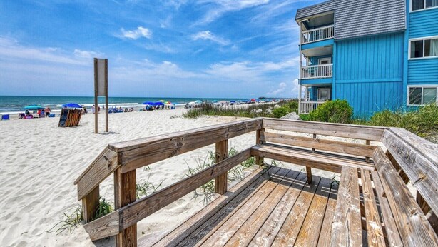 A beach with a wooden boardwalk, a blue building with balconies, and beach umbrellas. - Summer House - CANES, Three Bedroom, Two Bathroom, Second Row Community (Garden City Beach)