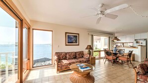 A spacious living area with a sofa, a round coffee table, and a dining set. A ceiling fan is visible, and there is a large window offering a view of the sea. - Oceanfront Kailua-Kona Townhome w/ Pool & Views! (Kailua-Kona)