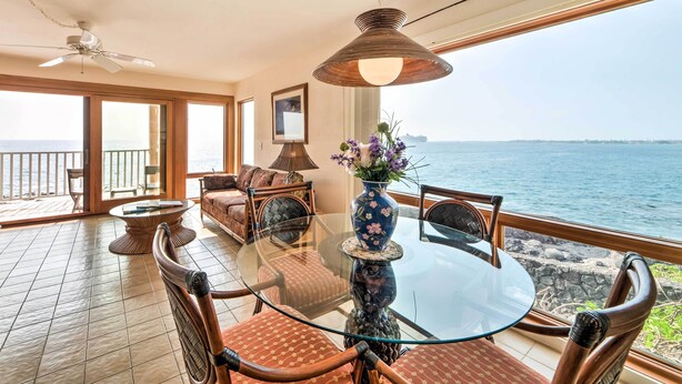 A spacious room with a glass table and chairs, a ceiling fan, a large window with a view of the sea, and a vase with flowers on the table. - Oceanfront Kailua-Kona Townhome w/ Pool & Views! (Kailua-Kona)