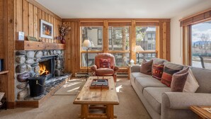 A cozy living room with a stone fireplace, wooden walls, a leather armchair, a sofa, and a view through large windows. - Nez Perce B1: Teton Village- Ground  Condo - Pool , Hot Tub Access (Teton Village)