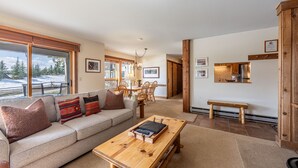 A spacious living room with a large window, a wooden coffee table, a sofa, and a dining area in the background. - Nez Perce B1: Teton Village- Ground  Condo - Pool , Hot Tub Access (Teton Village)