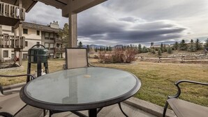 A covered patio with a glass table and chairs, overlooking a grassy area with a wooden fence and buildings in the background. - Nez Perce B1: Teton Village- Ground  Condo - Pool , Hot Tub Access (Teton Village)