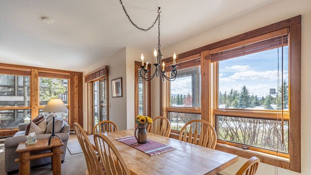 A dining area with a wooden table, chairs, and a chandelier. There is a sofa and a lamp in the adjacent living area. Large windows provide a view of trees and a building outside. - Nez Perce B1: Teton Village- Ground  Condo - Pool , Hot Tub Access (Teton Village)