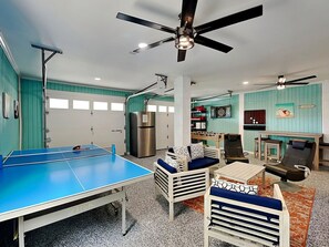 A room with a ping pong table, seating area, ceiling fan, and a dartboard.