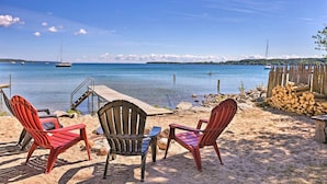 Una playa con tres sillas Adirondack, un muelle de madera y vistas al mar con barcos.