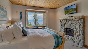 A bedroom with a stone fireplace, a bed with white linens, a window with a view of trees and mountains, and a television mounted on the wall.