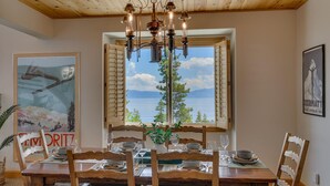 A dining area with a table set for four, wooden chairs, a chandelier, and a view of a lake through open shutters.
