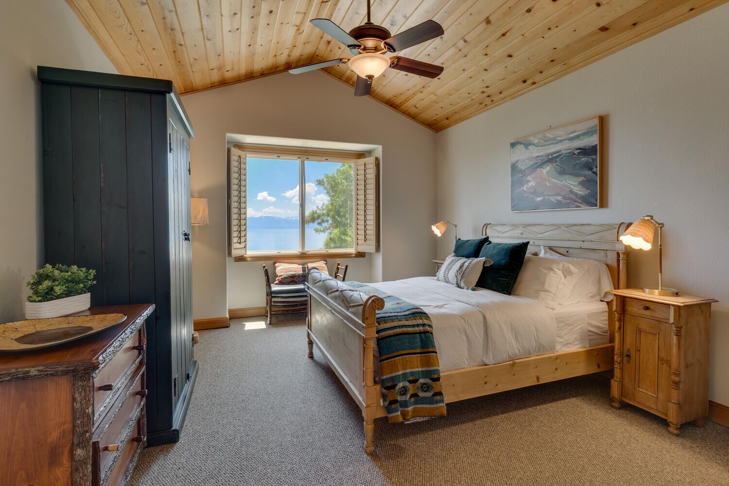 Quarto com teto de madeira, uma janela grande com vista para o exterior, cama com travesseiros e cobertor, mesa de cabeceira com abajur e cômoda com planta.