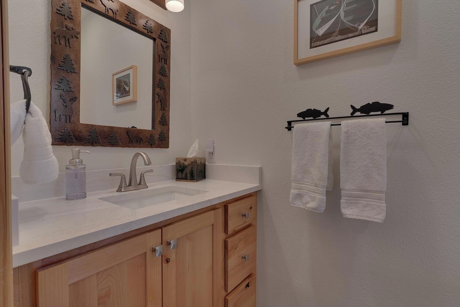 Un baño con tocador de madera, un espejo con marco decorativo, un lavabo, un perchero con dos toallas y un cuadro enmarcado en la pared.