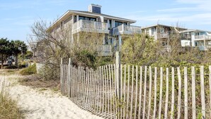 En fastighet som ligger vid stranden, med ett trästaket, en sandstrand och ett hus i flera våningar med balkonger.