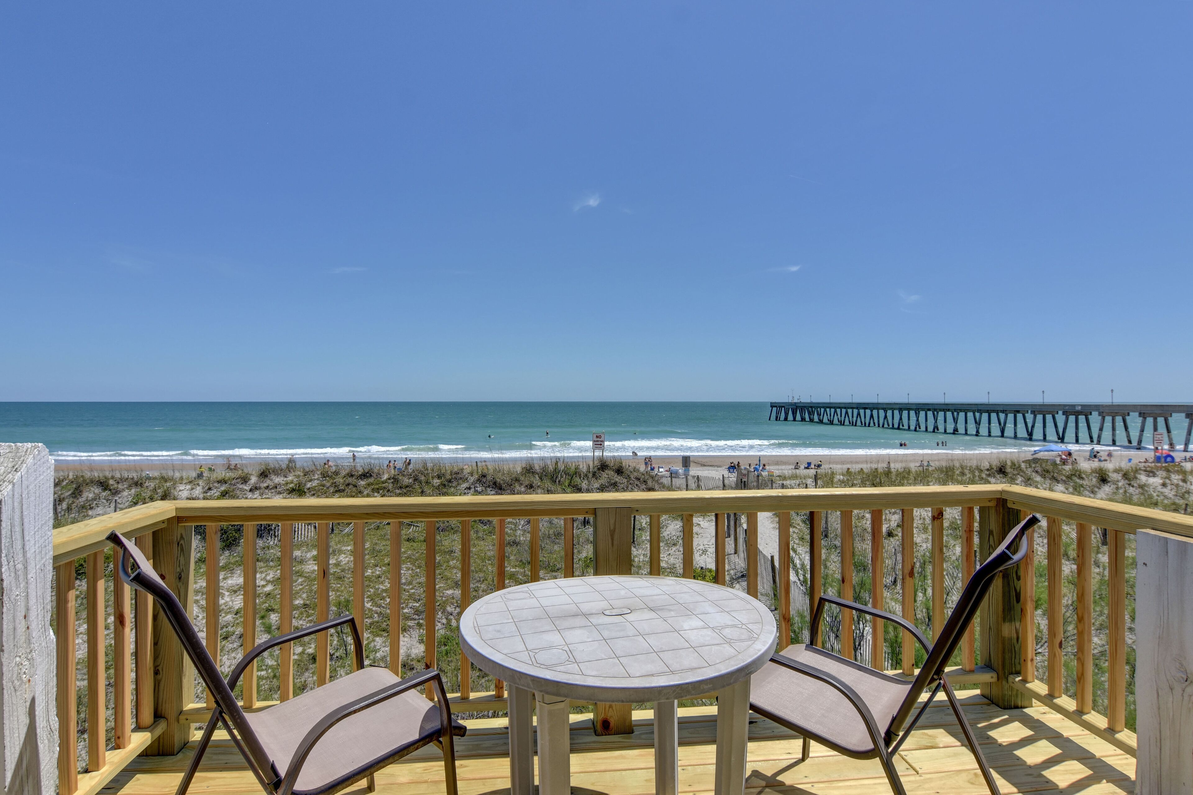 A wooden deck with a round table and two chairs, overlooking a beach and a pier.