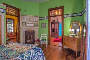 Une pièce comprenant un lit, une commode, une chaise et un miroir. On y trouve également une cheminée avec un manteau et une étagère où sont disposés des objets décoratifs.