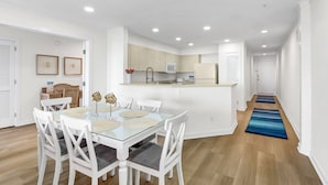 Una cocina moderna con armarios blancos, un microondas y un refrigerador. Hay una zona de comedor con una mesa de vidrio y sillas blancas.