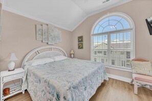 Quarto com uma cama grande, uma mesinha de cabeceira de vime branco, uma cadeira branca, uma janela grande em arco e obras de arte nas paredes.