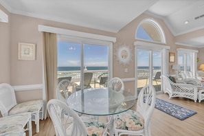 Una habitación con una mesa de vidrio rodeada de sillas de mimbre blanco, ventanas amplias con vista al océano y un adorno mural con diseño de sol.