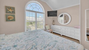 A bedroom with a large bed, a dresser, a TV, a window with blinds, and wall art. - Maultsby -Enjoy amazing ocean front views from this 10 bedroom home (Wrightsville Beach)