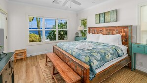 A bedroom with a wooden bed, a bench, a nightstand, a lamp, and a view of the outdoors through large windows. - Hale Ilikai By Parrish Kauai: fabulous family location with oceanfront views and (Koloa)