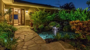Un chemin de pierres mène à une maison agrémentée d’une petite cascade et d’une végétation tropicale luxuriante.