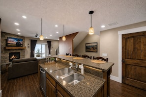A kitchen with granite countertops, a double sink, and a built-in oven. There is a fireplace, a dining area with a table and chairs, and a television mounted on the wall.