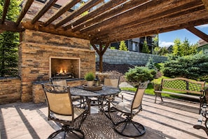 Une terrasse extérieure avec une cheminée en pierre, une table et des chaises en métal, et un banc en bois.