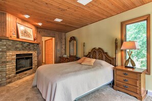 Ein Schlafzimmer mit Bett, Holz-Kopfteil, Nachttisch mit Lampe, Kommode, Kamin und großem Fenster.