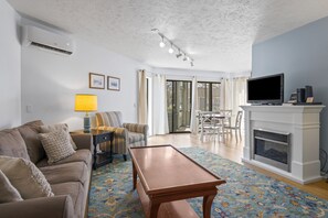 A living room with a sofa, a coffee table, a TV, a fireplace, and a dining area with chairs and a table. - Updated Condo | Dog Friendly | Downtown Charlevoix | Indoor/Outdoor Pool (Charlevoix)