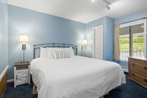 Une chambre à coucher comprenant un lit, des tables de chevet, une commode, une fenêtre avec des stores et une porte.