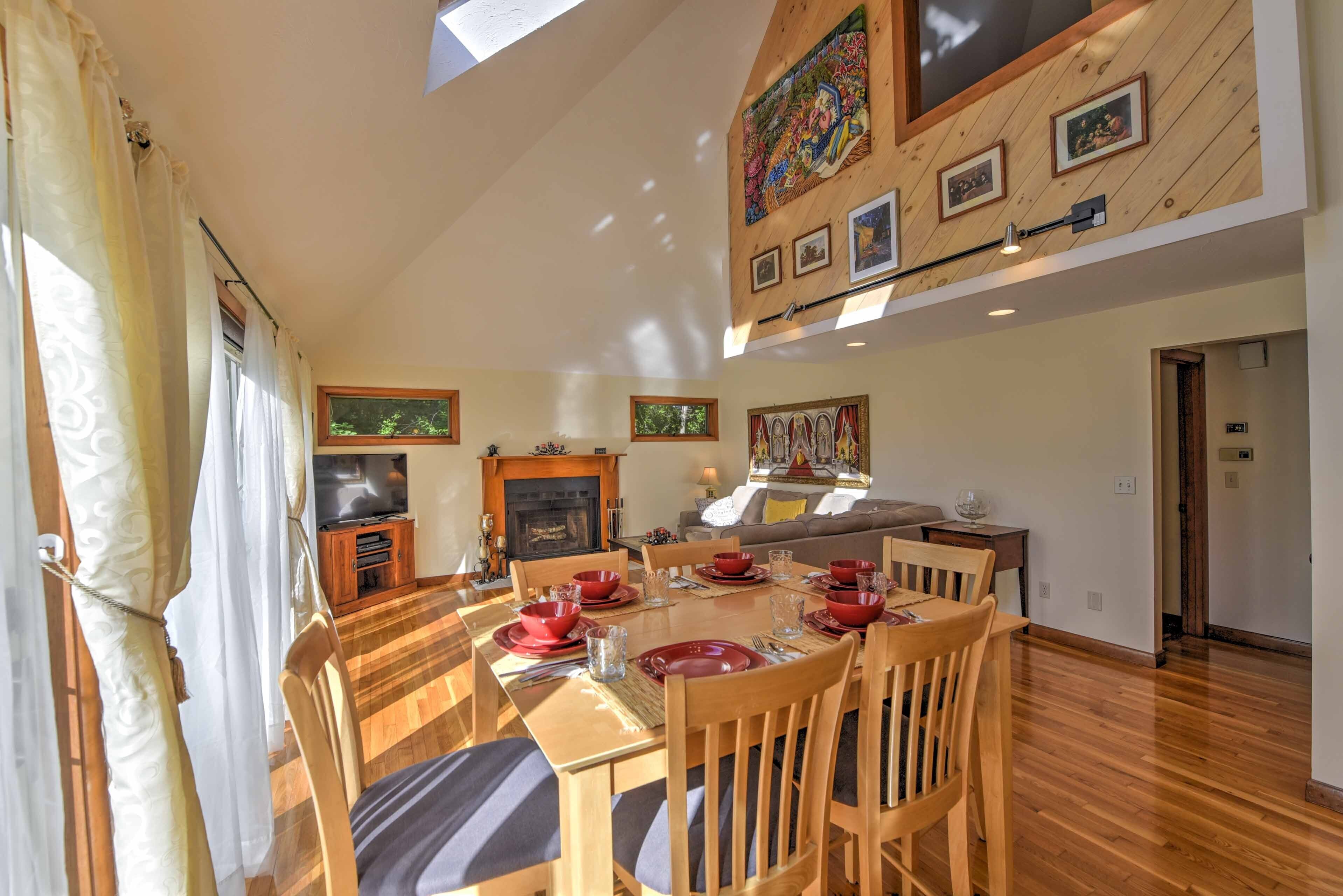 A dining area with a wooden table set for four, surrounded by chairs. A fireplace is visible in the background, and the room has wooden paneling on the walls.