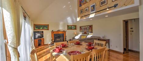 A dining area with a wooden table set for four, surrounded by chairs. A fireplace is visible in the background, and the room has wooden paneling on the walls.